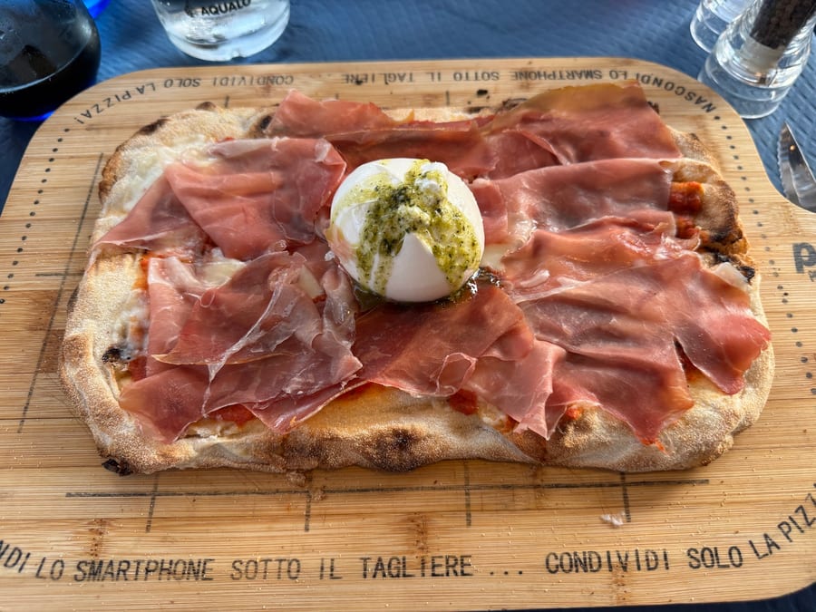 Pizza in teglia coperta di prosciutto crudo su tutta la superficie, con una burrata intera al centro e un cucchiaio di pesto sopra, servita sul taglie